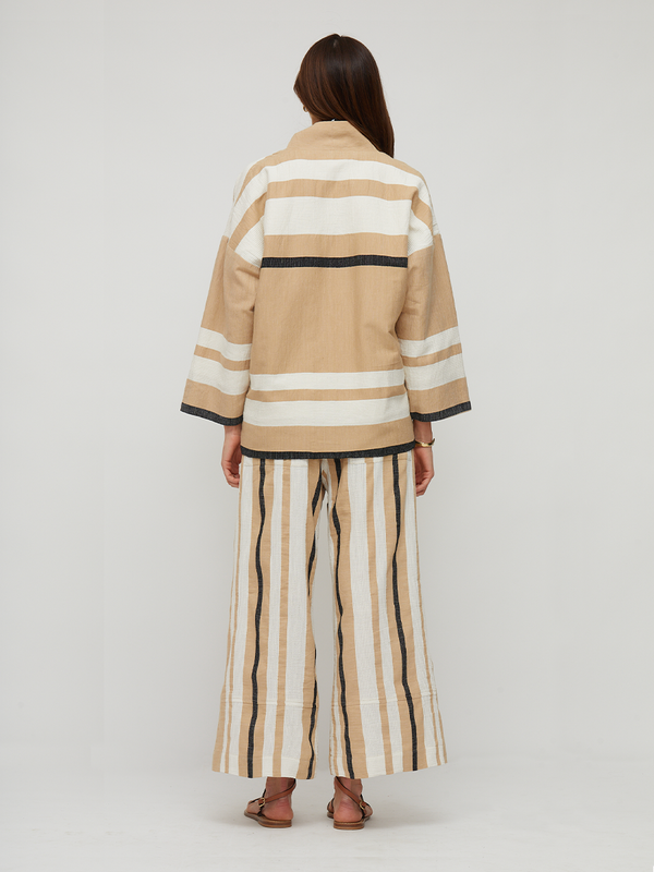 A model viewed from the back wearing a beige and white striped jacket with black trim and matching wide-leg striped pants, set against a plain light gray studio background.