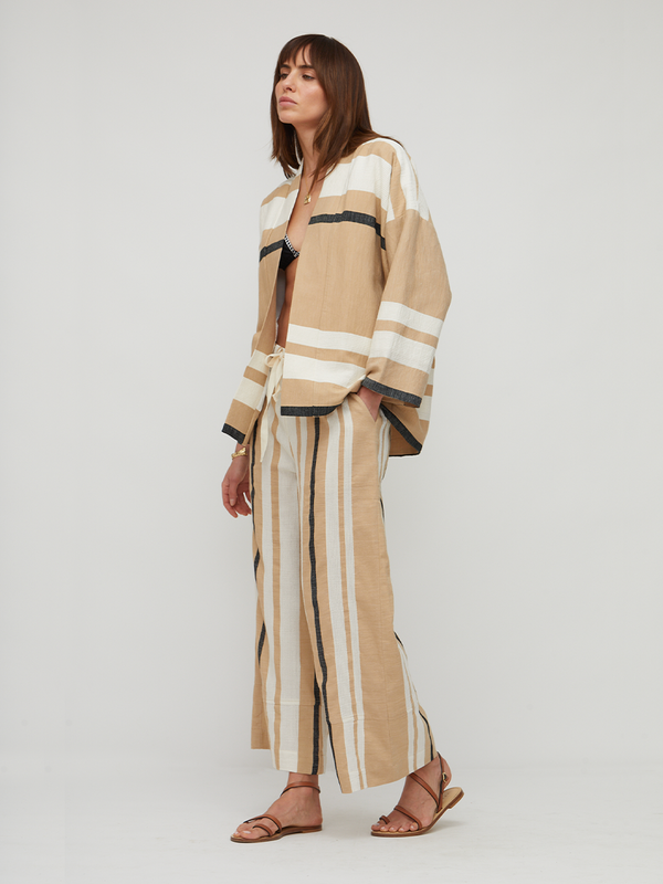 A woman models a matching beige and white striped linen two-piece set consisting of a cropped button-up shirt and wide-leg trousers, styled with brown leather sandals against a light gray studio background.