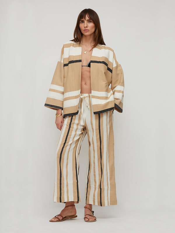 A model stands against a light gray studio background wearing a matching beige and cream striped lounge set consisting of a cropped jacket with black trim and wide-leg pants. She is also wearing brown leather slide sandals.