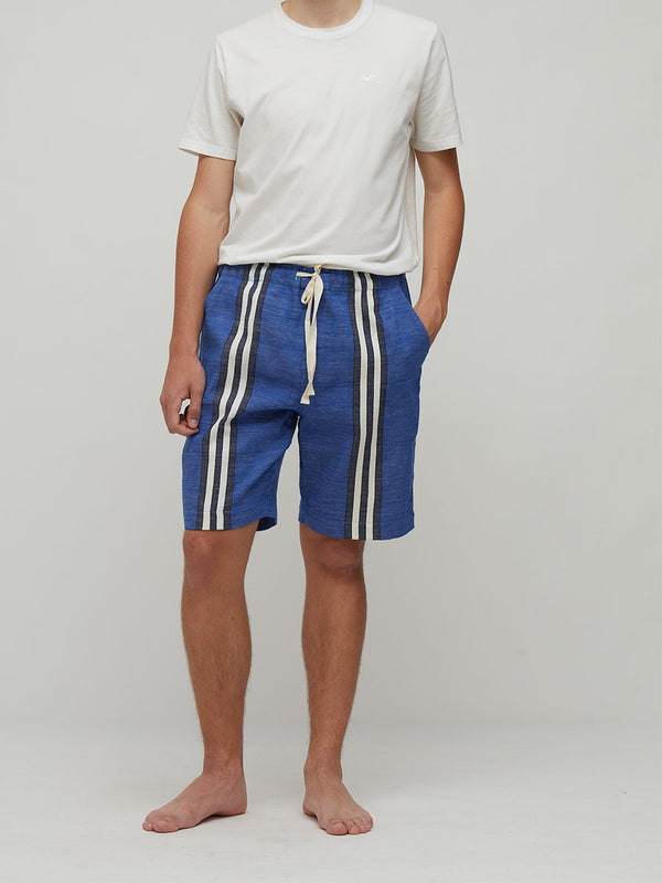 A man wearing lemlem handwoven  blue striped shorts with white side panels and a white drawstring, paired with a white t-shirt, standing against a light gray studio background.