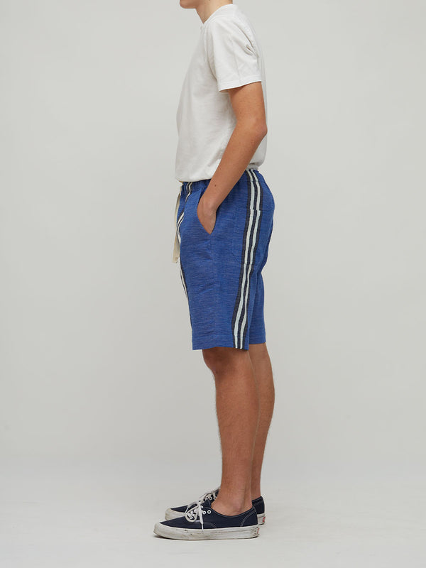 A side-profile studio shot of a model wearing lemlem handwoven blue shorts with white side stripes, a white t-shirt, and navy blue sneakers against a light gray background.