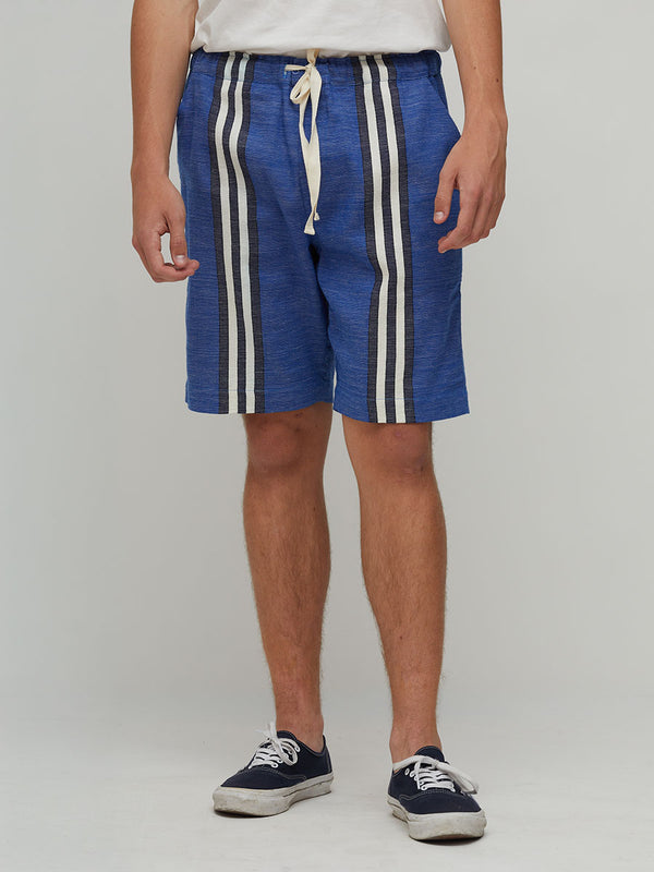 A person wearing lemlem handwoven  blue striped shorts with white side panels and a white drawstring, paired with a white top and navy sneakers, photographed against a light gray studio background.