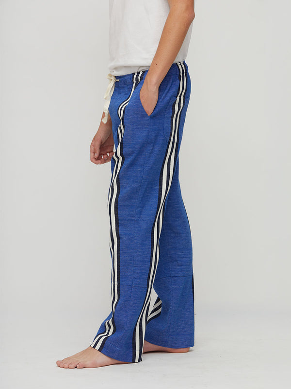 A person wearing loose handwoven lemlem blue pants with white and black side stripes, shown from the waist down against a plain light gray studio background.