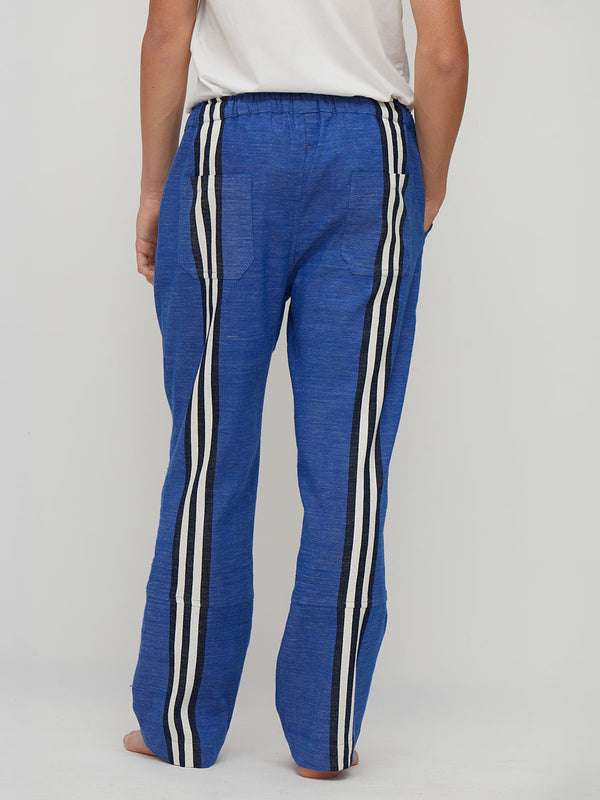 A person shown from the back wearing lemlem handwoven loose blue pants with two vertical white stripes bordered in black, paired with a white top against a light gray studio background.
