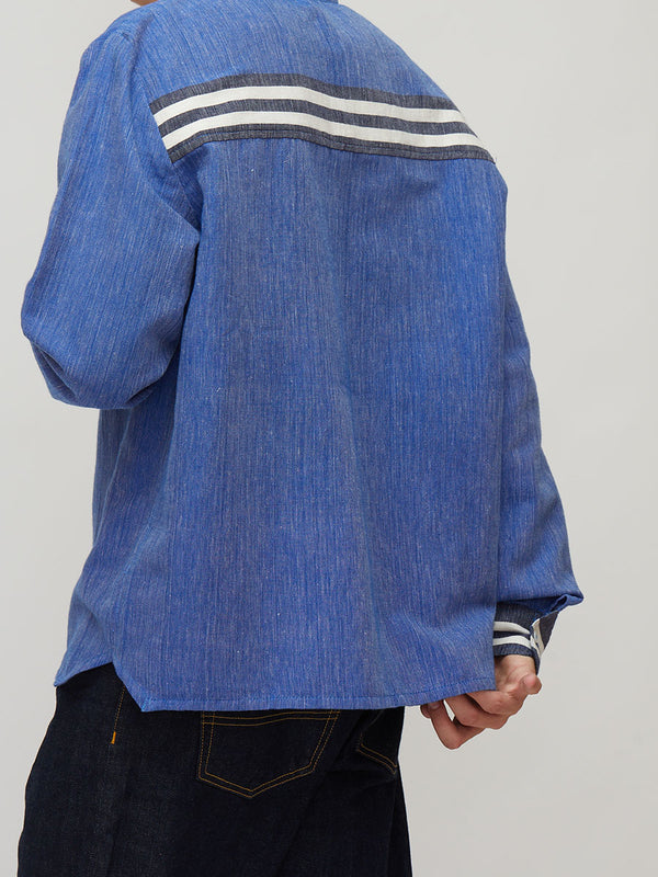 A person wearing a lemlem handwoven blue textured long-sleeve shirt with two horizontal white stripes across the upper back, paired with dark jeans, shown from a rear three-quarter angle against a light gray studio background.