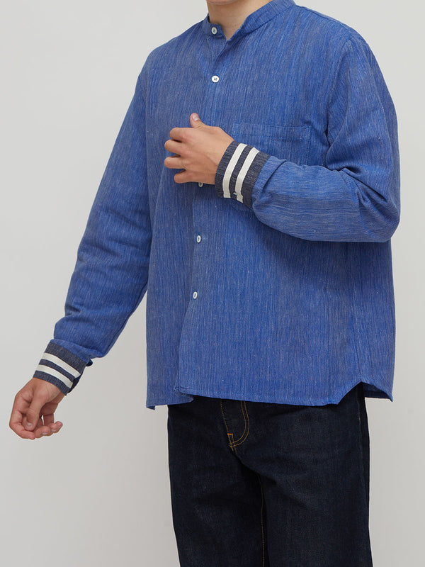 A man wearing lemlem handwoven blue textured long-sleeve button-up shirt with striped cuffs and dark jeans, shown from the neck down against a light gray background.