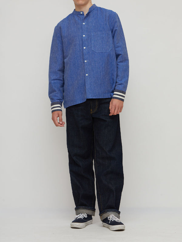 A model wearing lemlem handwoven  blue long-sleeve button-up shirt with striped cuffs, paired with dark blue jeans and navy sneakers, standing against a light gray studio background.