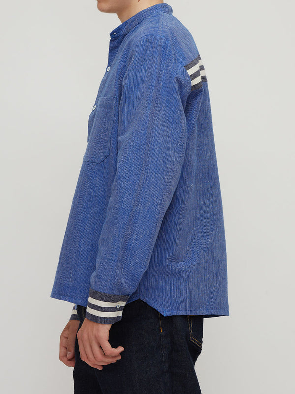 A side profile view of a person wearing a blue handwoven lemlem shirt with three white stripes on the sleeve and cuff, paired with dark jeans against a light gray background.