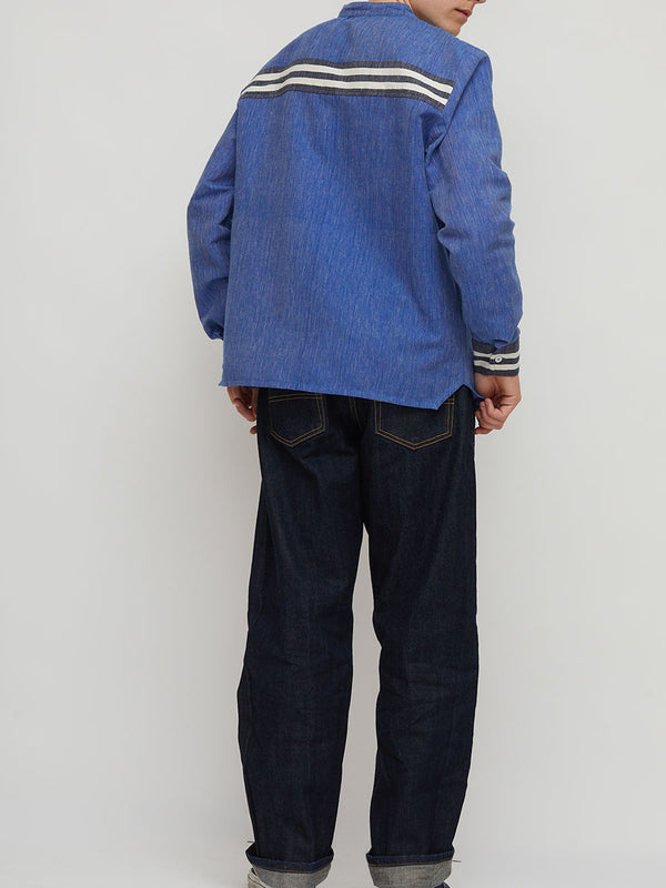A person shown from the back wearing a handwoven blue lemlem shirt featuring white and black stripes across the shoulders and cuffs, paired with dark jeans, against a plain light gray studio background.