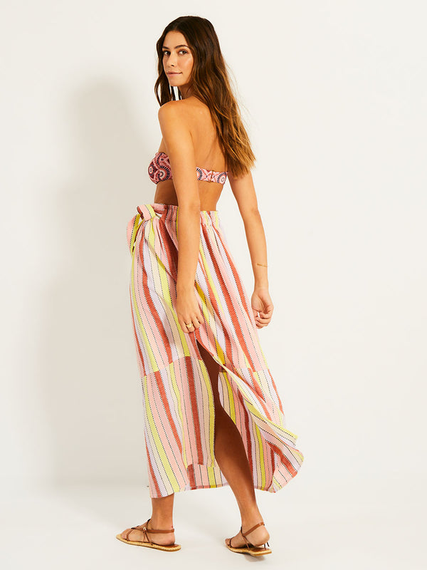 Back of a Woman Standing Wearing lemlem TOLA SKIRT  featuring soft sorbet-colored stripes in shades of peach, rust, and orchid, accented with a citrus yellow pop.

