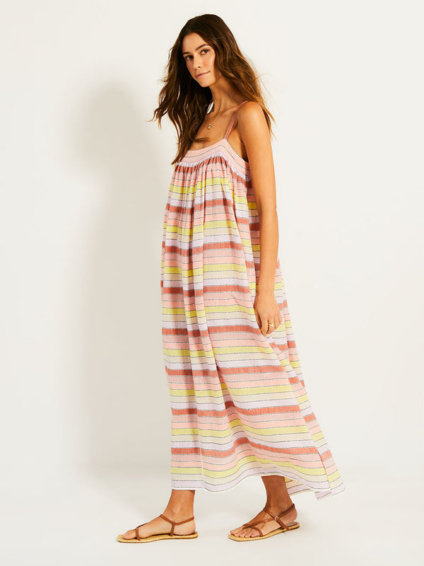 Side Shot of a Woman Standing Wearing lemlem EDA Slip Dress featuring soft sorbet-colored stripes in shades of peach, rust, and orchid, accented with a citrus yellow pop.
