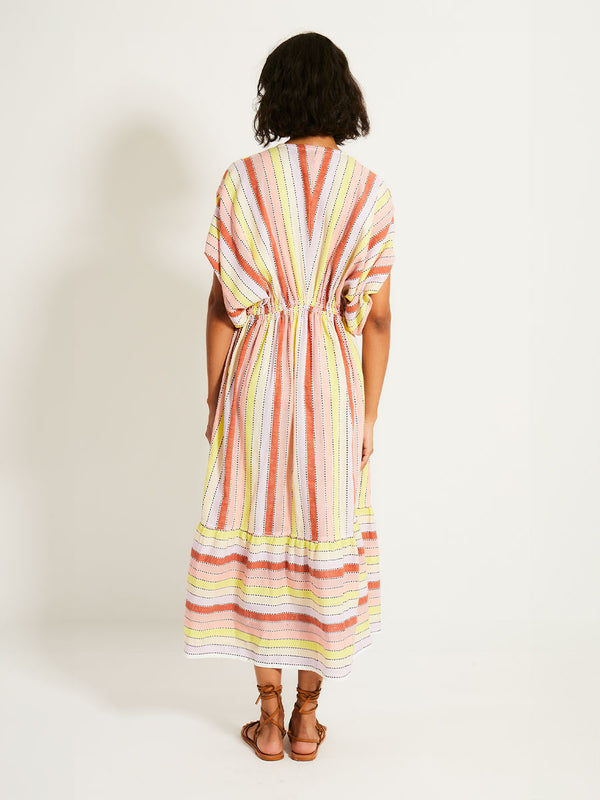 Back of a Woman Standing Wearing lemlem LEILA Plunge Dress featuring soft sorbet-colored stripes in shades of peach, rust, and orchid, accented with a citrus yellow pop.
