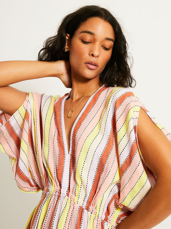 Close up on a Woman Standing Wearing lemlem LEILA Plunge Dress featuring soft sorbet-colored stripes in shades of peach, rust, and orchid, accented with a citrus yellow pop.
