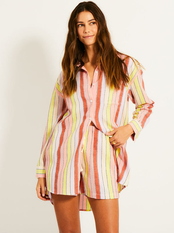 Woman Standing Wearing lemlem MARIAM Shirt  featuring soft sorbet-colored stripes in shades of peach, rust, and orchid, accented with a citrus yellow pop.

