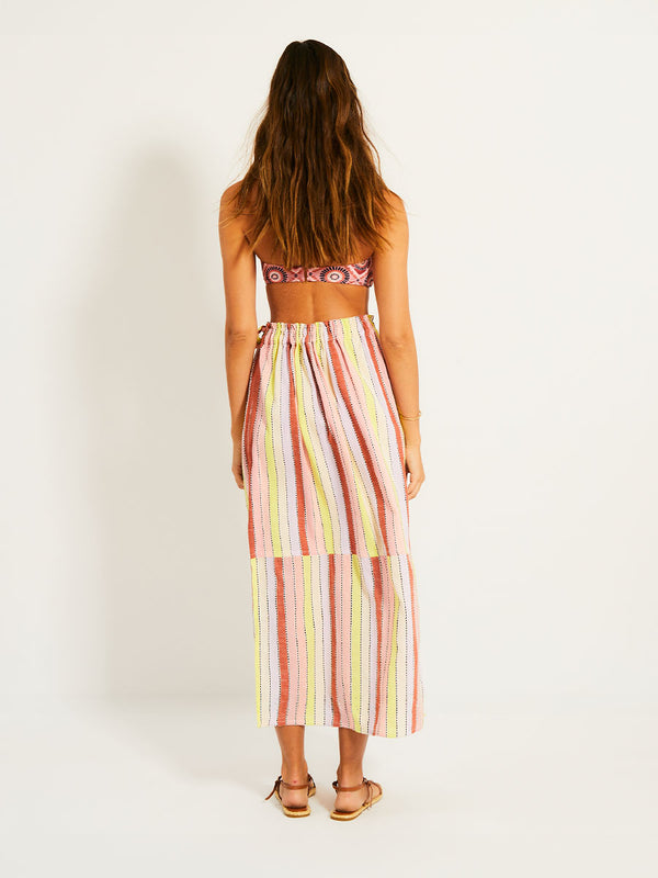 Back of a Woman Standing Wearing lemlem TOLA SKIRT  featuring soft sorbet-colored stripes in shades of peach, rust, and orchid, accented with a citrus yellow pop.
