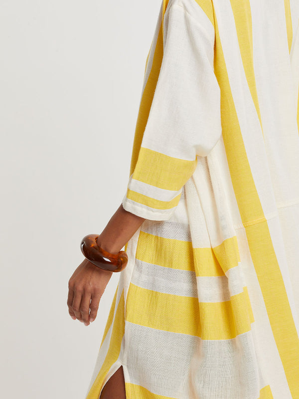 Woman standing wearing lemlem handwoven FANA Caftan featuring a wide block stripe pattern in rich yellow on a creamy vanilla background.