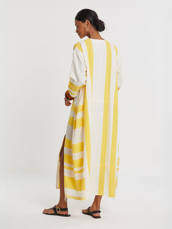 Woman standing wearing lemlem handwoven FANA Caftan featuring a wide block stripe pattern in rich yellow on a creamy vanilla background.