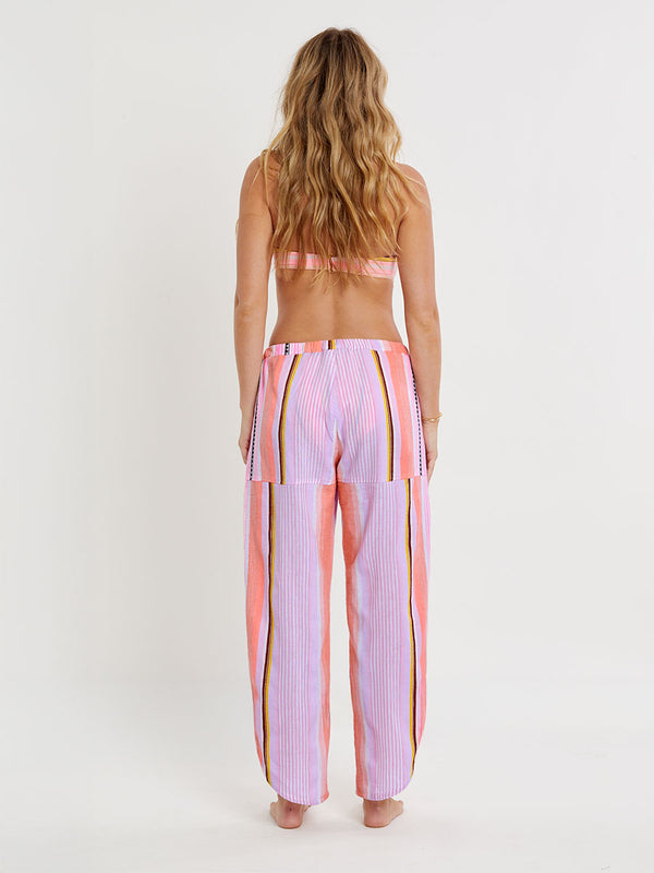 Woman standing wearing lemlem handwoven MIMI Flyaway pants featuring intricate stripe pattern interlaced with delicate mini Tibeb detailing, expressed in a soft yet vivid palette of cool pink, warm tangerine, rose, orchid, and accents of peach and deep burgundy.