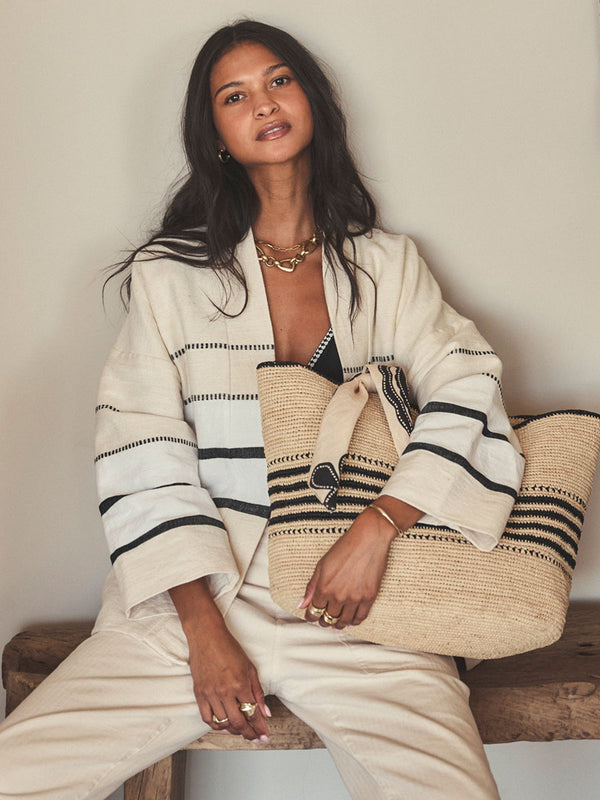 Woman holding a woven bag, wearing a white outfit with black stripes, sitting on a wooden bench.