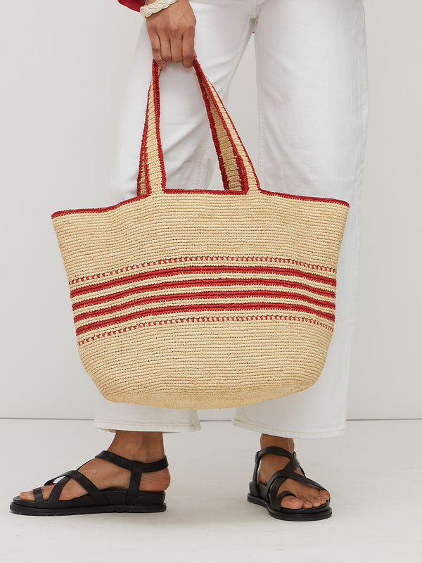 Woman holding a lemlem MIRA Raffia tote bag with red stripes on a beige background