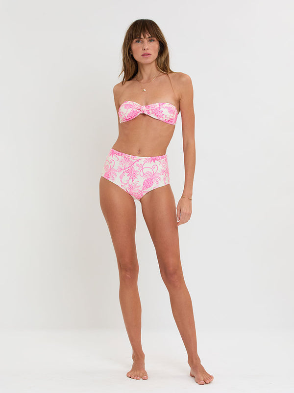 Woman standing wearing lemlem AVA Bandeau Top and ELSI High Waist Bottom featuring a unique custom tropical pineapple motif in vibrant fuchsia on an ivory background.