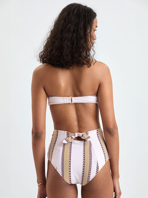 back of a Woman Standing wearing lemlem AVA Bandeau Top and ELSI High Waist Bottom featuring detailed bands of deep burgundy, bronze, and pink stripes beautifully placed on a light cream 