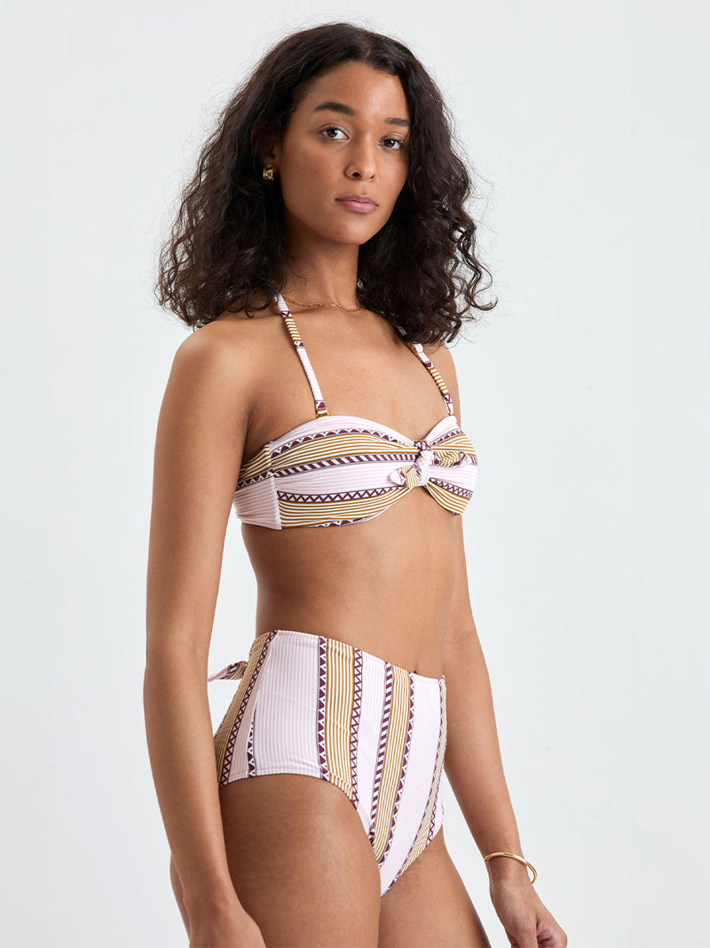 Woman Standing wearing lemlem AVA Bandeau Top and ELSI High Waist Bottom featuring detailed bands of deep burgundy, bronze, and pink stripes beautifully placed on a light cream 