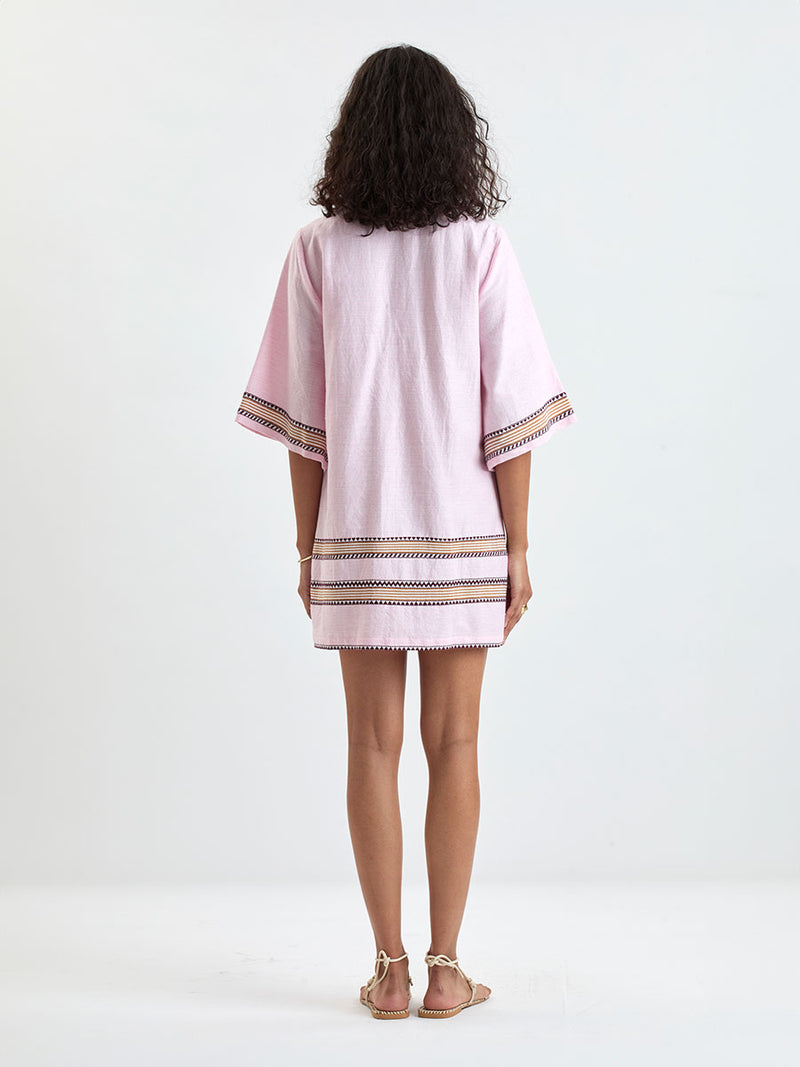 back of a Woman Standing wearing lemlem BELKIS Caftan featuring detailed bands of deep burgundy, bronze, and pink stripes beautifully placed on a light cream 