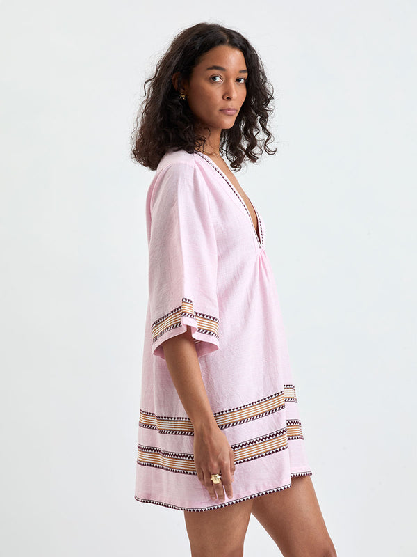 side shot of a Woman Standing wearing lemlem BELKIS Caftan featuring detailed bands of deep burgundy, bronze, and pink stripes beautifully placed on a light cream 