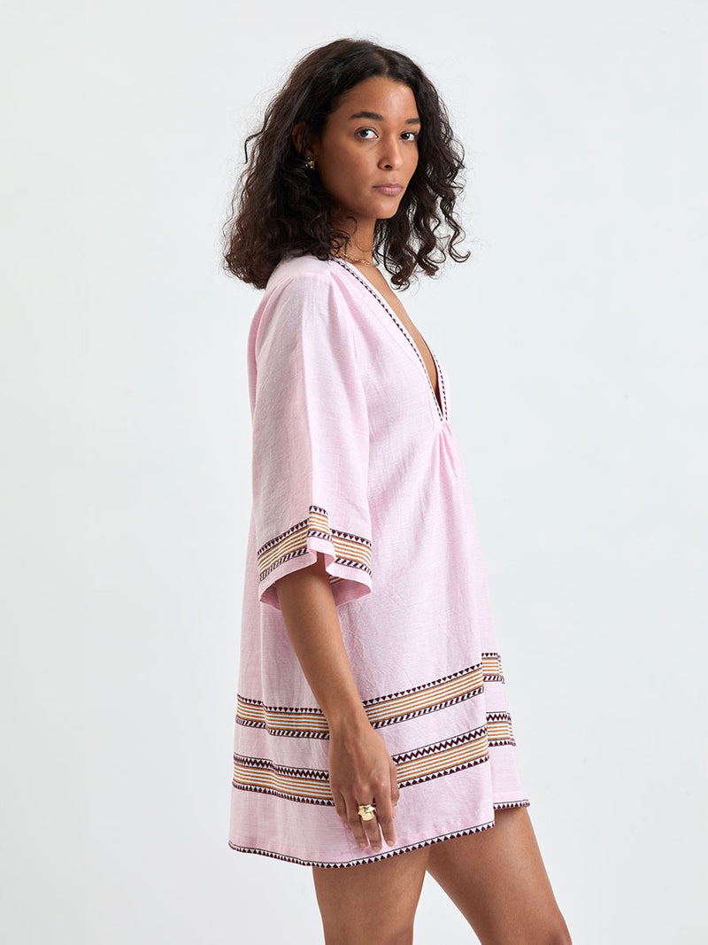 side shot of a Woman Standing wearing lemlem BELKIS Caftan featuring detailed bands of deep burgundy, bronze, and pink stripes beautifully placed on a light cream 