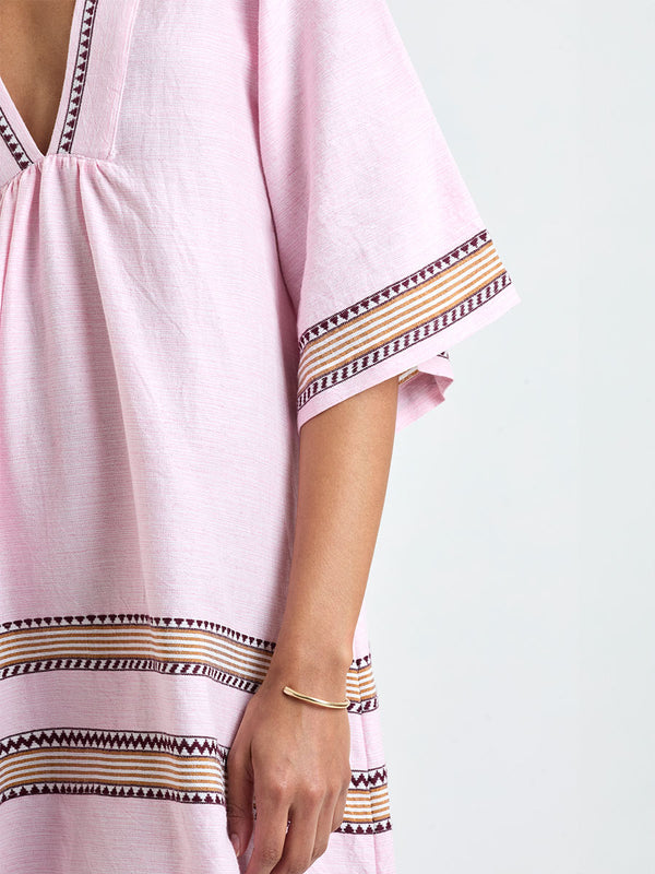 close up on a Woman Standing wearing lemlem BELKIS Caftan featuring detailed bands of deep burgundy, bronze, and pink stripes beautifully placed on a light cream 