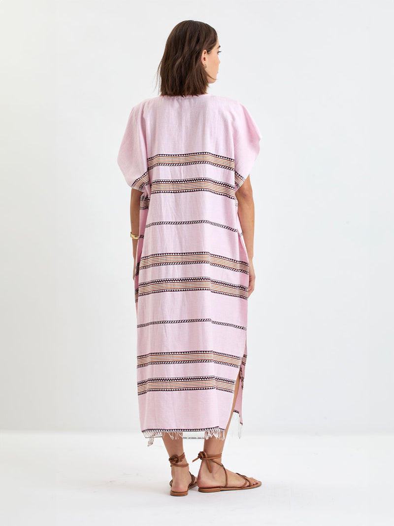 Back of a Woman Standing wearing lemlem handwoven DALILA Caftan featuring detailed bands of deep burgundy, bronze, and pink stripes beautifully placed on a light cream 