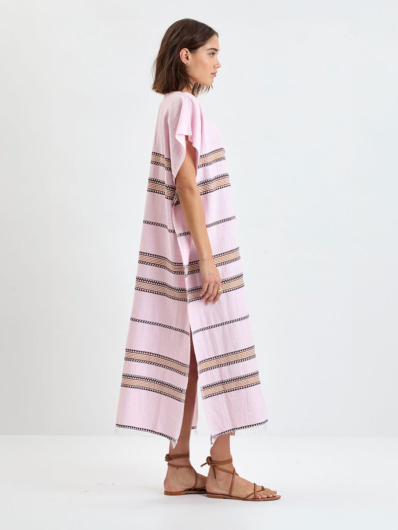 Woman Standing wearing lemlem handwoven DALILA Caftan featuring detailed bands of deep burgundy, bronze, and pink stripes beautifully placed on a light cream 