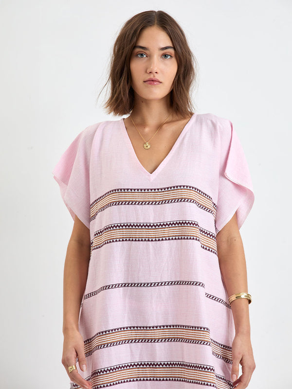Woman Standing wearing lemlem handwoven DALILA Caftan featuring detailed bands of deep burgundy, bronze, and pink stripes beautifully placed on a light cream 