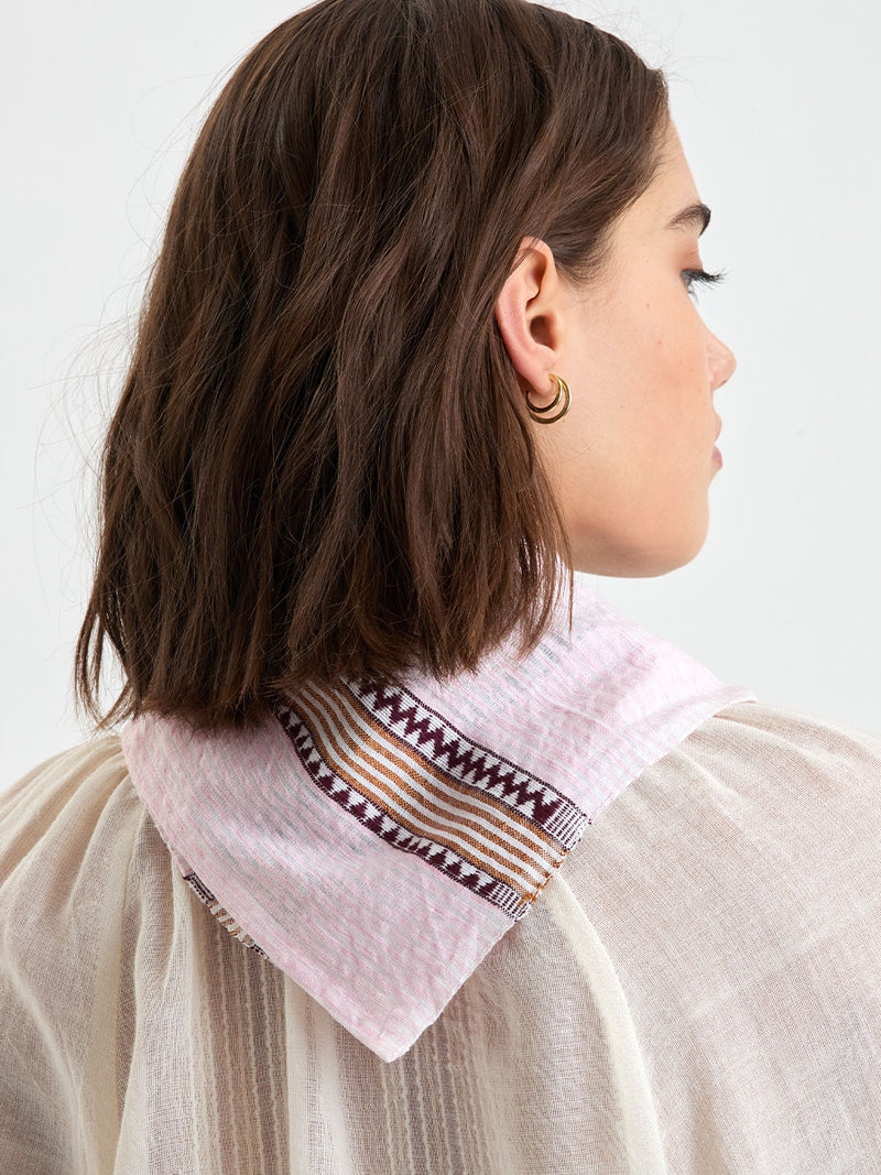 Back close up of a woman standing wearing lemlem DANA Bandana featuring detailed bands of deep burgundy, bronze, and pink stripes beautifully placed on a light cream 