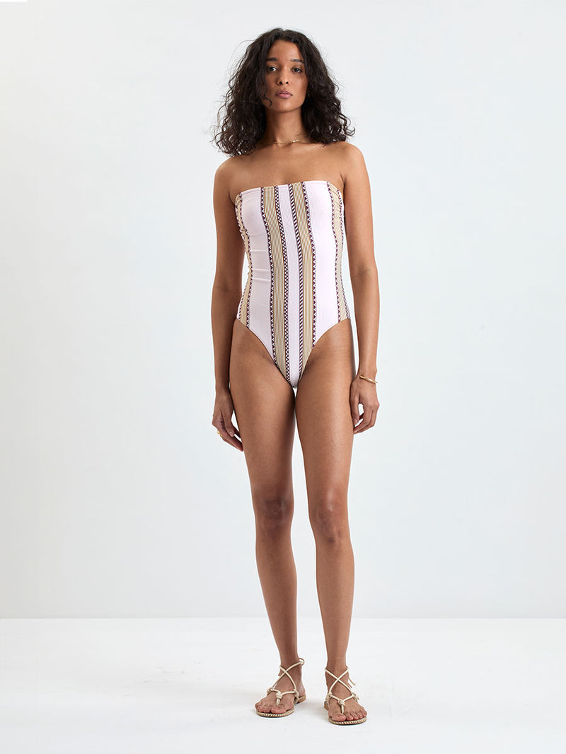 Woman Standing wearing lemlem DINHA One Piece featuring detailed bands of deep burgundy, bronze, and pink stripes beautifully placed on a light cream 