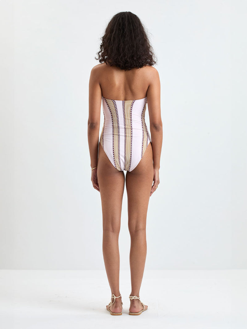 back of a Woman Standing wearing lemlem DINHA One Piece featuring detailed bands of deep burgundy, bronze, and pink stripes beautifully placed on a light cream 