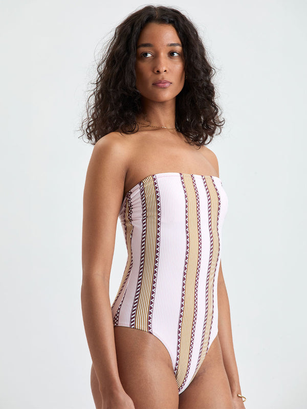 Woman Standing wearing lemlem DINHA One Piece featuring detailed bands of deep burgundy, bronze, and pink stripes beautifully placed on a light cream