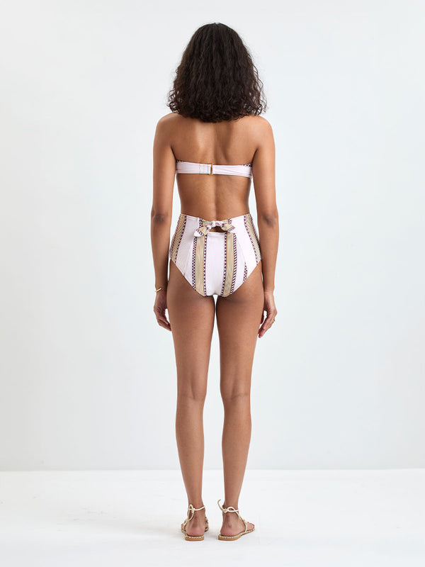 back of a Woman Standing wearing lemlem AVA Bandeau Top and ELSI High Waist Bottom featuring detailed bands of deep burgundy, bronze, and pink stripes beautifully placed on a light cream 