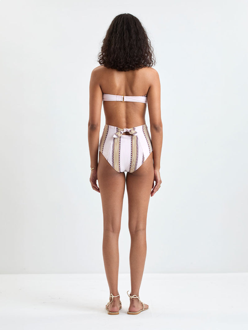 back of a Woman Standing wearing lemlem AVA Bandeau Top and ELSI High Waist Bottom featuring detailed bands of deep burgundy, bronze, and pink stripes beautifully placed on a light cream 
