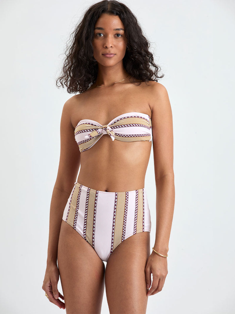 Woman Standing wearing lemlem AVA Bandeau Top and ELSI High Waist Bottom featuring detailed bands of deep burgundy, bronze, and pink stripes beautifully placed on a light cream 