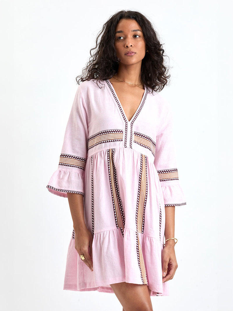 Woman Standing wearing lemlem handwoven HANNA Flutter Dress featuring detailed bands of deep burgundy, bronze, and pink stripes beautifully placed on a light cream 