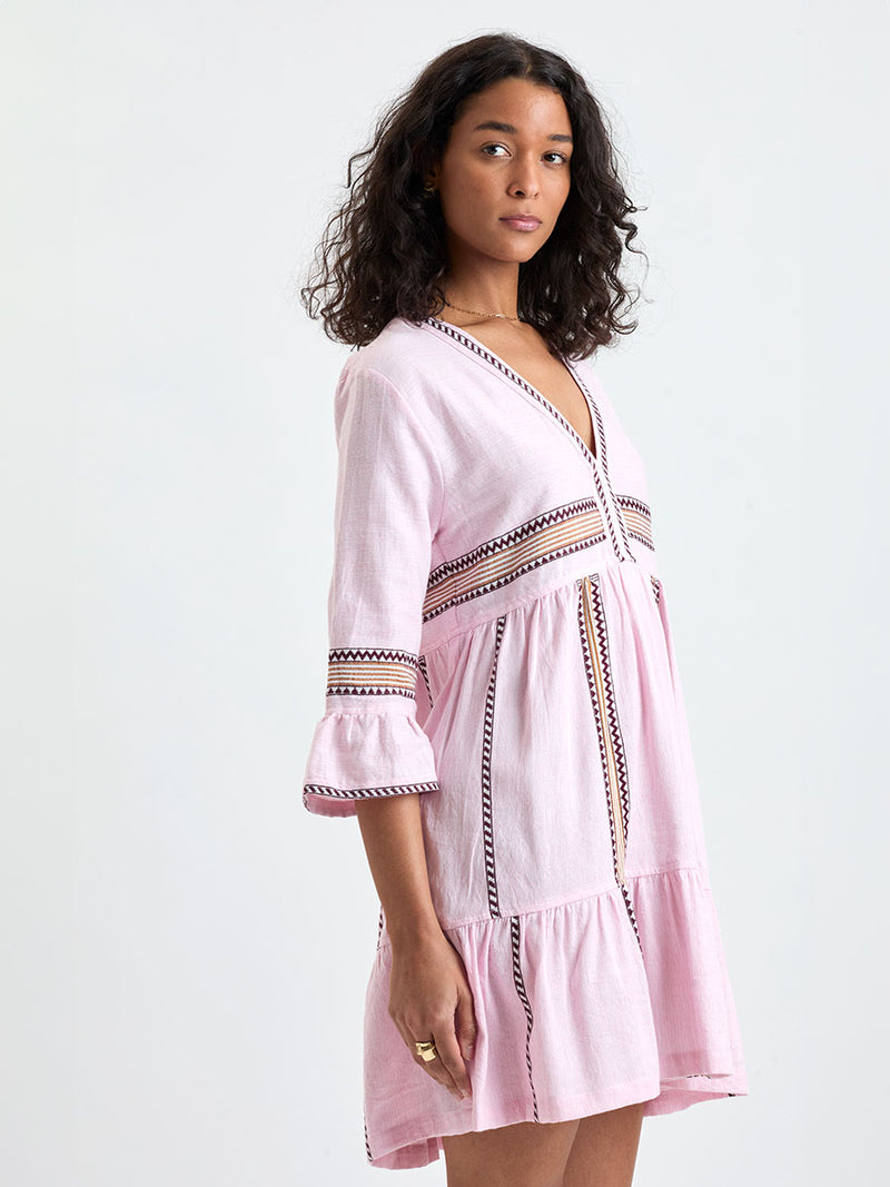 Woman Standing wearing lemlem handwoven HANNA Flutter Dress featuring detailed bands of deep burgundy, bronze, and pink stripes beautifully placed on a light cream 