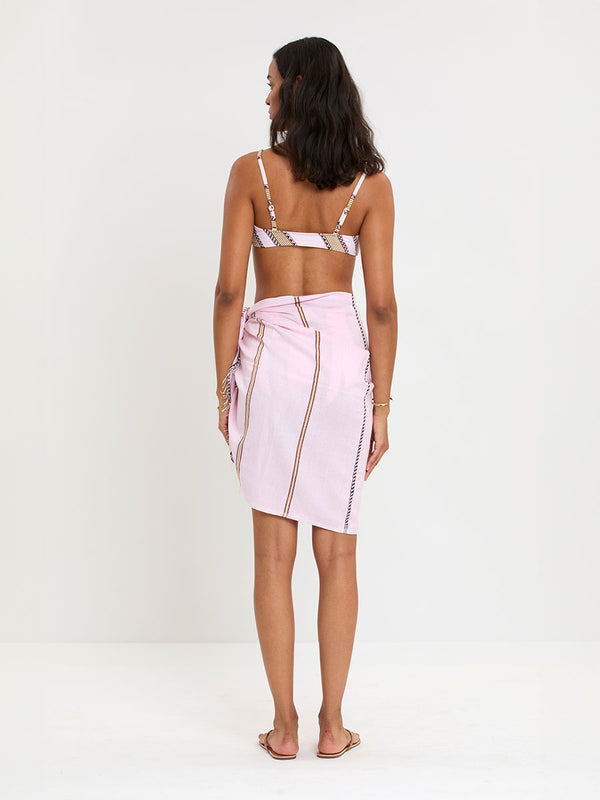 Back of a Woman Standing wearing lemlem LEMA Sarong featuring detailed bands of deep burgundy, bronze, and pink stripes beautifully placed on a light cream 
