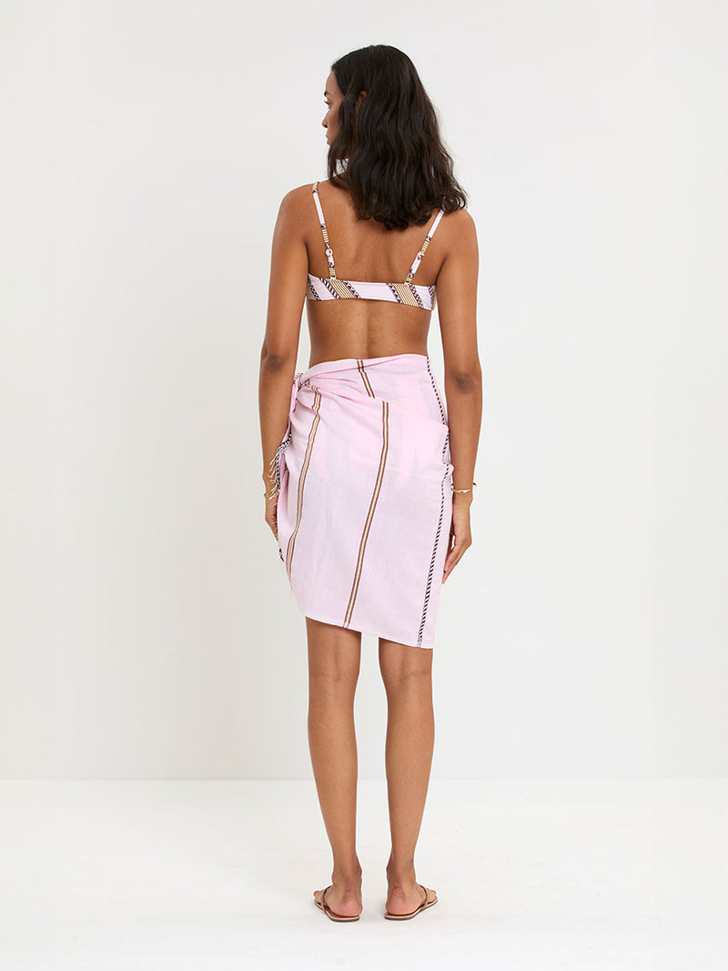 Back of a Woman Standing wearing lemlem LEMA Sarong featuring detailed bands of deep burgundy, bronze, and pink stripes beautifully placed on a light cream 