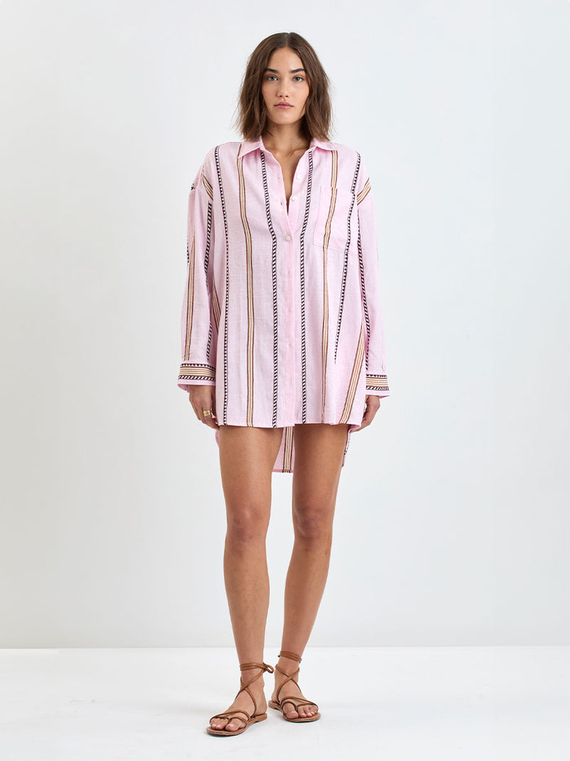 \Woman Standing wearing lemlem MARIAM Shirt featuring detailed bands of deep burgundy, bronze, and pink stripes beautifully placed on a light cream 