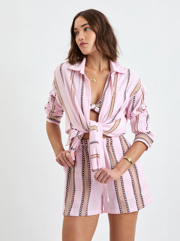 Woman Standing wearing lemlem MARIAM Shirt featuring detailed bands of deep burgundy, bronze, and pink stripes beautifully placed on a light cream 