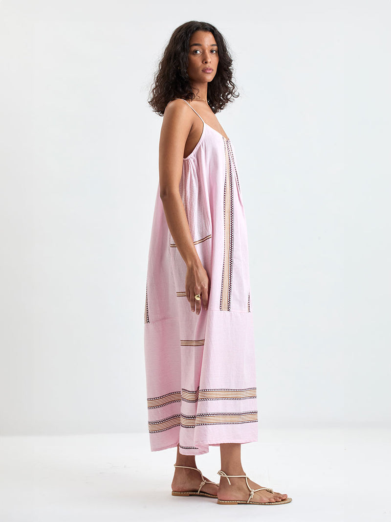 Woman Standing wearing lemlem NIA Slip Dress featuring detailed bands of deep burgundy, bronze, and pink stripes beautifully placed on a light cream 
