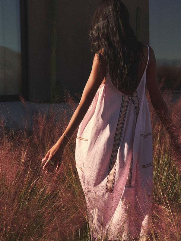 Back of a Woman standing wearing lemlem handwoven slip dress featuring pink color and tibeb pattern in brown color