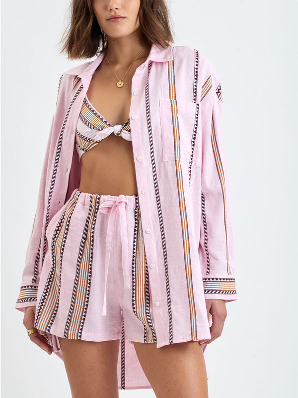 close up on a Woman Standing wearing lemlem MARIAM Shirt featuring detailed bands of deep burgundy, bronze, and pink stripes beautifully placed on a light cream 